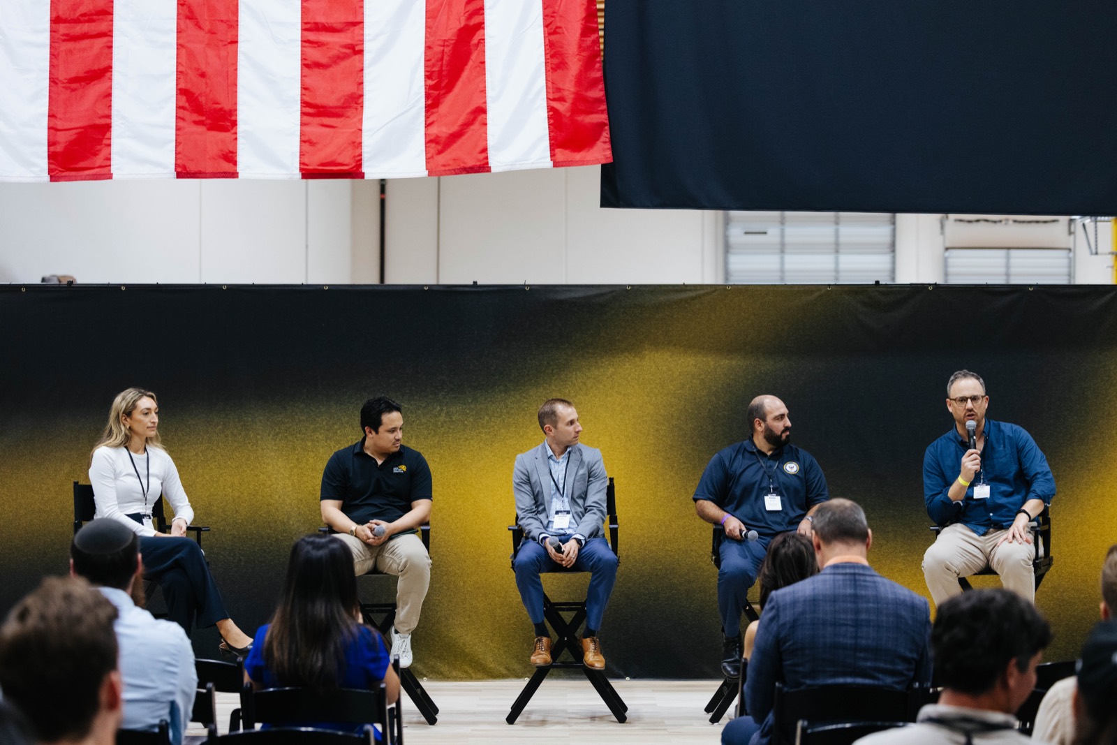 Five panelists seated on stage under an American flag, audience in the foreground